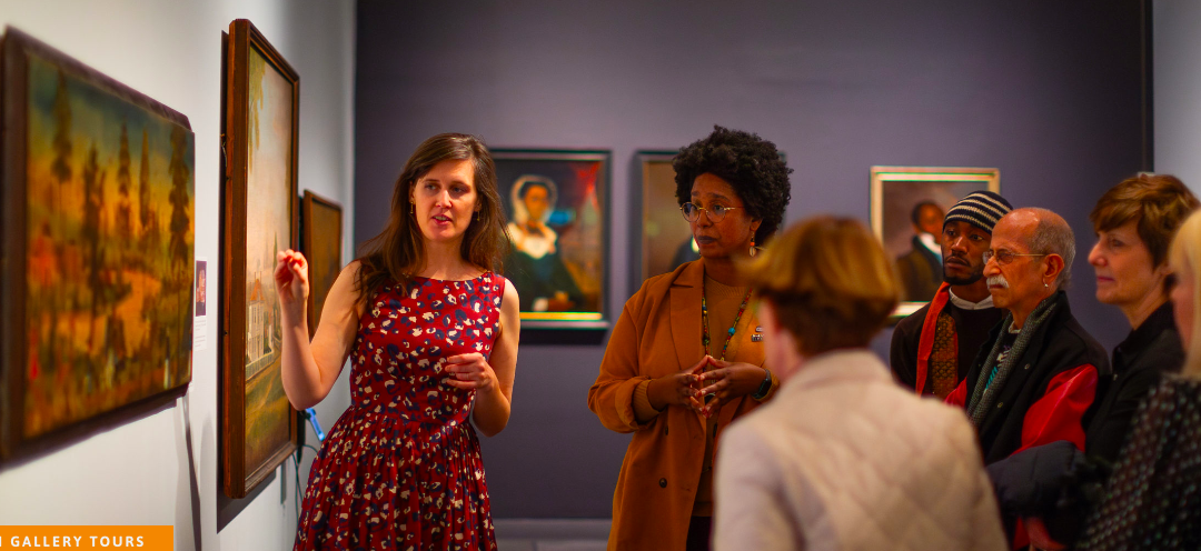 Woman points to a framed artwork in front of a group of adults in museum gallery.