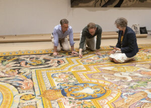 Three adults crouch over a yellow French carpet to study weave and design.